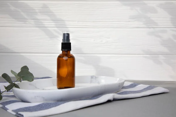 Amber bottle with facial cosmetics on a tray with a branch of eucalyptus on the gray wooden background, facial serum. Front view