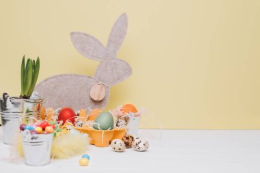 Beyaz ahşap arka planda gri tavşanlı sarı karton tepside çok renkli yumurtalar. Paskalya arkaplanı, sembol. Ön görünüm