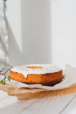 Yuvarlak Paskalya keki, beyaz protein kremi ve beyaz arka planda genç ağaç dallarıyla süslenmiş renkli şekerlerle süslenmiş. Ön görünüm