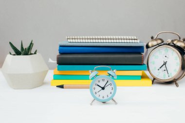 Kırtasiyeli işyeri. Çok renkli not defterleri, mavi alarm saati, gri bir duvara karşı beyaz ahşap bir masada sulu. Ön görünüm