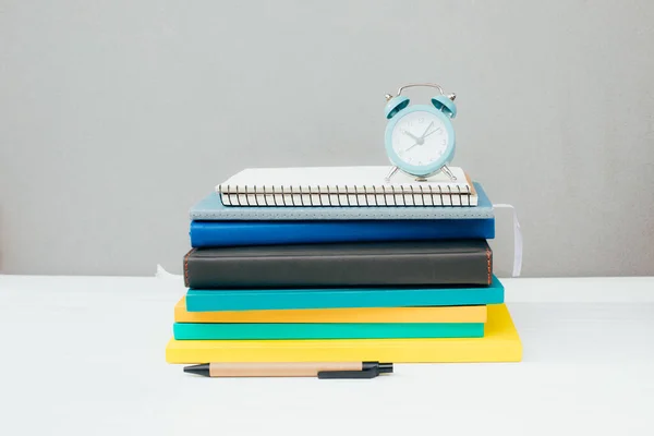 stock image Workplace with stationery. Multicolored notepads, blue alarm clock, succulent on a white wooden table against a gray wall. Front view