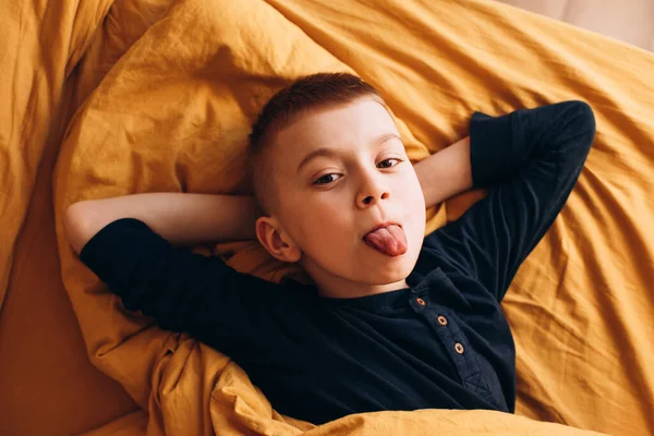A joyful boy lies in the yellow bad and shows tongue. The boy in his bad in the morning. Front view