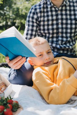 Aile pikniği. Baba ve oğlu yeşil bir bahçede, güneşli bir havada piknik yapıyor, pizza yiyor ve kitap okuyor. Ön görünüm