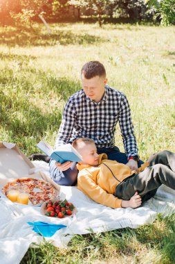 Aile pikniği. Baba ve oğlu yeşil bir bahçede, güneşli bir havada piknik yapıyor, pizza yiyor ve kitap okuyor. Ön görünüm