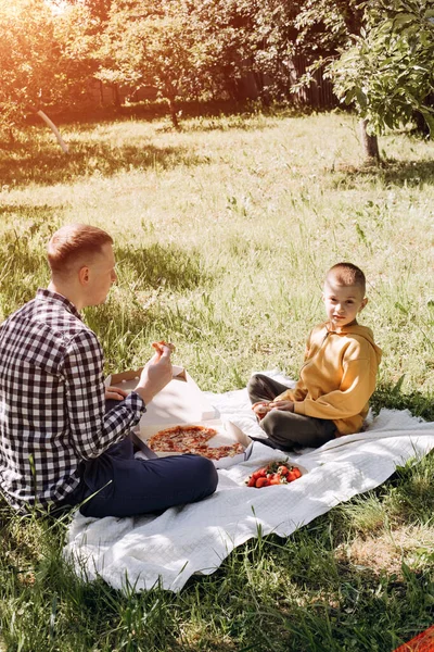 stock image Family picnic. Dad and son in a green garden in sunny weather on a picnic eating pizza. Front view