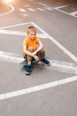Çocuklar için açık hava aktiviteleri. Parlak elbiseli neşeli genç bir çocuk kaykayın üzerinde oturuyor. Beyaz bir okul çocuğu yürüyüşe çıktı. Üst görünüm