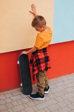 Çocuklar için açık hava aktiviteleri. Renkli bir duvarın arkasında parlak elbiseli, kaykaylı neşeli bir genç. Beyaz bir okul çocuğu yürüyüşe çıktı. Ön görünüm