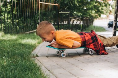 Çocuklar için açık hava aktiviteleri. Parlak elbiseli neşeli genç bir çocuk kaykayın üzerinde yatıyor. Beyaz bir okul çocuğu yürüyüşe çıktı. Ön görünüm