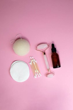 Çevre dostu cihazlar. Pembe kuvars yüz masajı silindiri, yüz serumu, kulak çubukları ve pembe arka planda yeniden kullanılabilir pamuk pedleri. Düz yatıyordu
