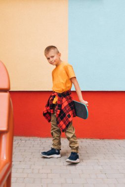 Çocuklar için açık hava aktiviteleri. Parlak elbiseli neşeli genç bir çocuk renkli bir duvarın arkasında kaykay tutuyor. Beyaz bir okul çocuğu yürüyüşe çıktı. Ön görünüm