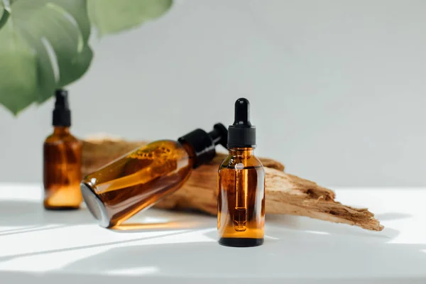 stock image Amber bottles with facial treatment on a wooden stand on a white table with green leaves on backdrop. Front view