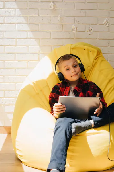 A school-age boy in black headphones sits on a yellow bean bag with a tablet in his hands. Children and gadgets. Front view