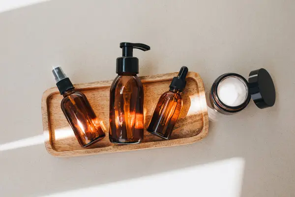 Amber bottles with facial cosmetics on a wooden tray on the beige concrete background with a streak of daylight. Flat lay