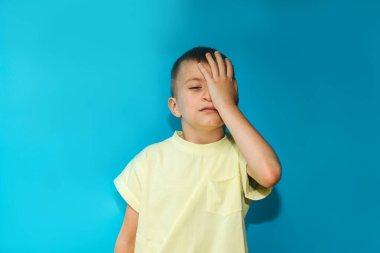 A 6-7-year-old boy in a yellow T-shirt shows a gesture of being bored covering his face with one hand on a blue background. Close-up portrait, front view