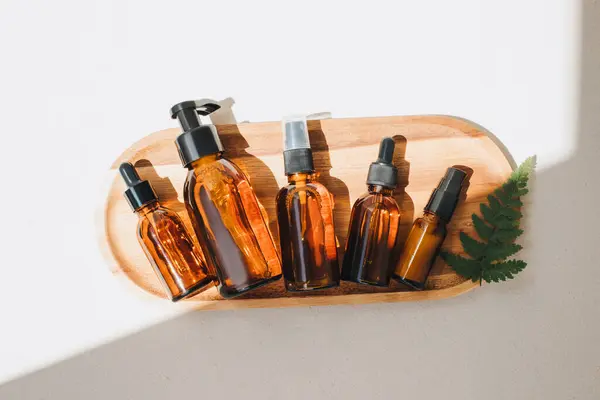 Amber bottles with facial cosmetics on a wooden tray on the beige concrete background with a streak of daylight. Flat lay