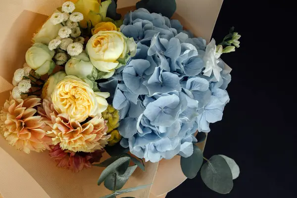 Bouquet of flowers on a black background. Floral background, postcard. The bouquet with blue hydrangea and yellow roses is packed in light pink paper. Top view