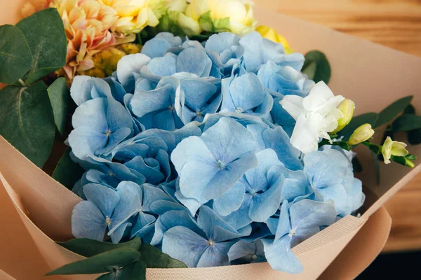 Bouquet of flowers on a wooden background. Floral background, postcard. The bouquet with blue hydrangea and yellow roses is packed in light pink paper. Front view