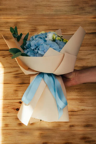 Bouquet of flowers on a wooden background. Floral background, postcard. The bouquet with blue hydrangea and yellow roses wrapped in light pink paper in the hand. Front view