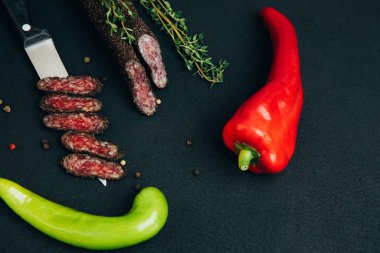Acı biber kağıtları ve kekik ile mutfak bıçağında sosis. Siyah beton arka planda füme sosis. Üst görünüm