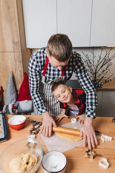 Dad and son at a wooden table in the kitchen are going to make cookies by rolling out the dough with a wooden rolling pin. Front view