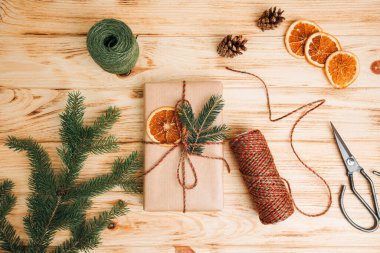 Noel arkaplanı. Kahverengi kağıda sarılmış kuru turuncu ve kozalaklı ağaç dallarıyla birlikte açık ahşap zemin üzerinde. Noel hediyesi paketleme süreci. Üst görünüm