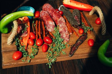 Yeşil kağıt ve domatesli ahşap tepside farklı tip ve boyutlarda sosisler. Koyu ahşap arka planda tütsülenmiş sosis seti. Üst görünüm