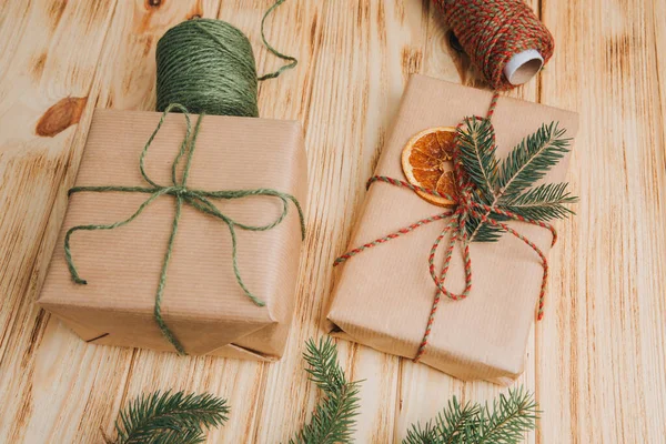 Christmas background. Gift wrapping in brown paper with dry orange and branch of coniferous tree on a light wooden background. The process of packing a Christmas gift. Top view