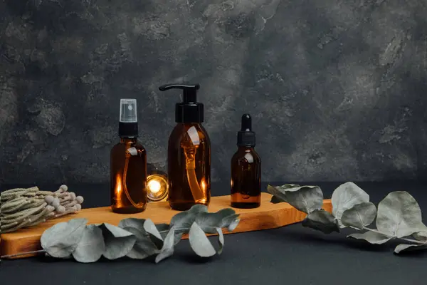 Amber bottles with facial cosmetics on a wooden tray with a dry branches on a dark background, facial serum. Front view