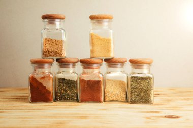 Hafif ahşap arka planda cam kavanozlarda bir grup baharat. Paprika, otlar, hardal, sarımsak, ön manzara, seçici odak