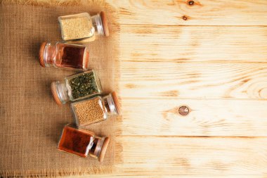 Hafif ahşap arka planda cam kavanozlarda bir grup baharat. Paprika, otlar, hardal, sarımsak. Üst görünüm, seçici odak