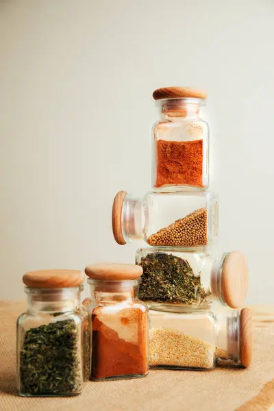 A group of seasonings in glass jars on a light wooden background. Paprika, herbs, mustard, garlic, front view, selective focus