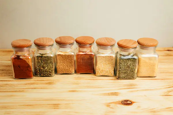 A group of seasonings in glass jars on a light wooden background. Paprika, herbs, mustard, garlic. Top view, selective focus