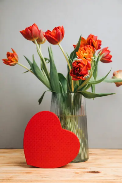 Red bouquet of tulips in a vase with red gift box in the shape of heart. Background with a bouquet of flowers. Valentines day gifts. Front view