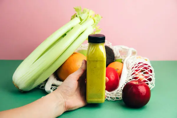 Fruit and vegetable smoothies in transparent plastic bottles. Green smoothie in the hand on a pink-green background with avocado, apples and celery. The concept of healthy eating. Front view
