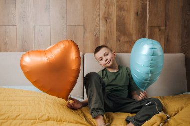 Yeşil tişörtlü, turuncu ve mavi balonlu, kötünün üstünde kalp şeklinde bir çocuk. Sevgililer Günü. Bir çocuk portresi. Ön görünüm