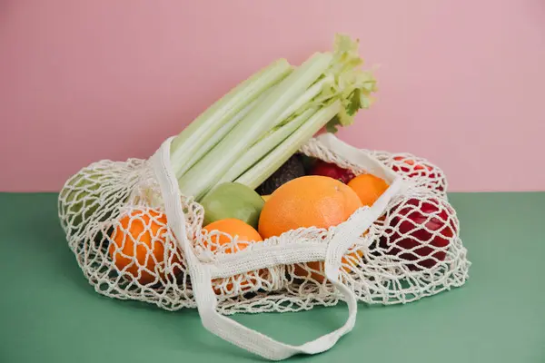 Fruit and vegetable in beige mesh bag. Apple, orange, avocado on a pink-green background. The concept of healthy eating, shopping. Front view