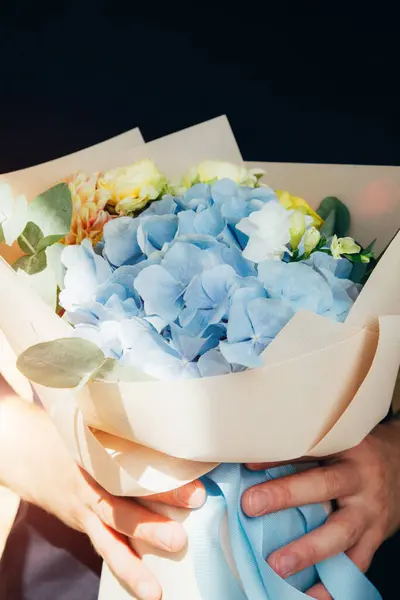 Floral background, postcard. The bouquet with blue hydrangea and yellow roses wrapped in light pink paper in the hand. Front view
