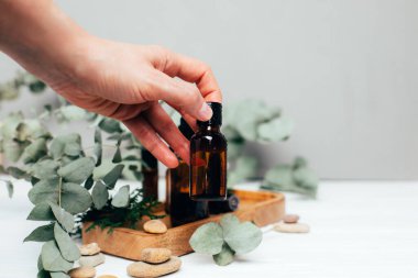 Kadın eli, gri ahşap masada bir okaliptüs dalı, yüz temizleyici ve serum olan bir kehribar şişesini bir tepside tutuyor. Ön görünüm