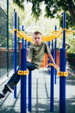Yeşil tişörtlü okul çağındaki bir çocuk güneşli havada oyun parkında, yatay barlarda vakit geçirir. Ön görünüm