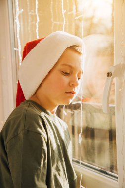 A boy in red Christmas hat near the window in bright sunlight. Children and pets. Portrait of a child. Christmas eve. Front view clipart