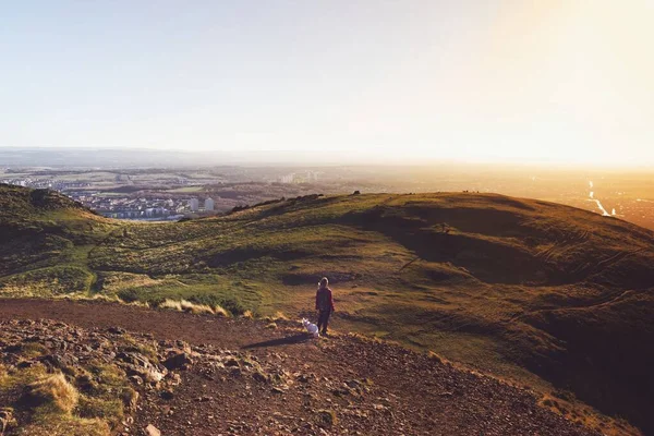 Experience the simple joys of life in this uplifting photograph taken at Arthur Seat, one of Edinburghs natural treasures. A person and her West Highland White Terrier, also known as a Westie, are