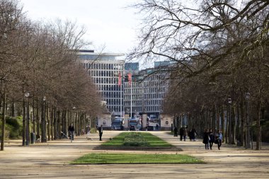 Görüntü, uzaktaki çağdaş bir binaya doğru uzanan yapraksız ağaçlarla çevrili bir şehir parkının içinden bir cadde gösteriyor. İnsanlar yolda yürürken, eğlenirken görülürler.