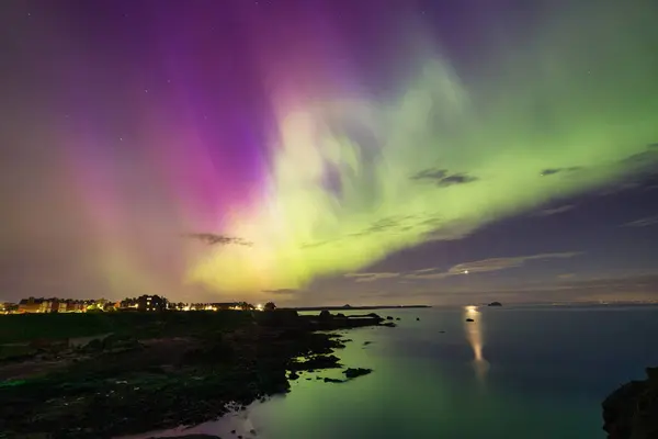 stock image An enchanting image of the Aurora Borealis lighting up the night sky over the calm waters of North Berwick, Scotland. The vivid green and purple lights stretch across the sky, reflecting beautifully