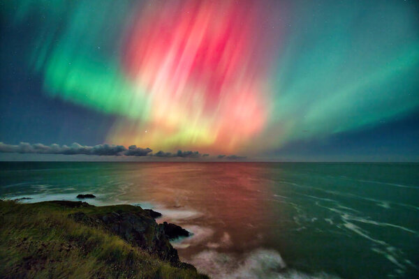A stunning Aurora Borealis display lights up the sky over the cliffs of St. Abbs, UK. The vibrant green and pink hues of the northern lights contrast beautifully with the dark ocean and rugged