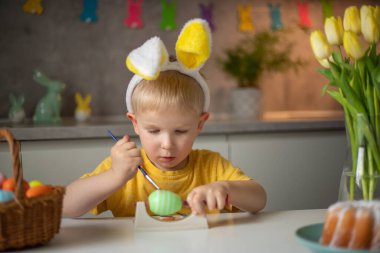 A little boy wearing bunny ears prepares painted Easter eggs for Easter while sitting at the kitchen table. Happy easter.