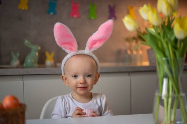 happy cute little baby girl wearing bunny ears on easter day sitting at the table in the kitchen.