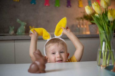 A cute little boy wearing bunny ears on Easter day wants to eat a chocolate easter bunny. A child plays egg hunt for Easter. Charming child celebrates Easter at home.
