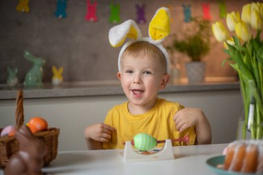 Emotional portrait of a cheerful little boy wearing bunny ears on Easter day who laughs merrily, plays with colorful Easter eggs sitting at a table in the kitchen.