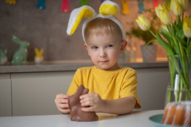 A cute little boy wearing bunny ears on Easter day is eating a chocolate Easter bunny. A child plays egg hunt for Easter. Charming child celebrates Easter at home.