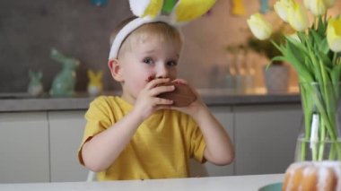 Little cute boy wearing bunny ears eating chocolate easter bunny while sitting at table at home.